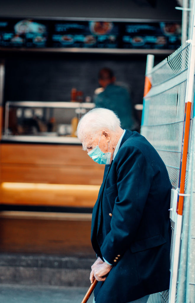 Une personne âgée portant le masque appuyée sur un mur regardant le sol