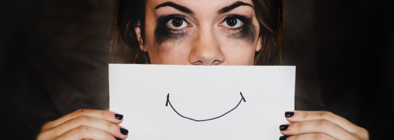 Une femme dont le maquillage a coulé cache le bas de son visage avec une feuille sur laquelle est dessiné un sourire