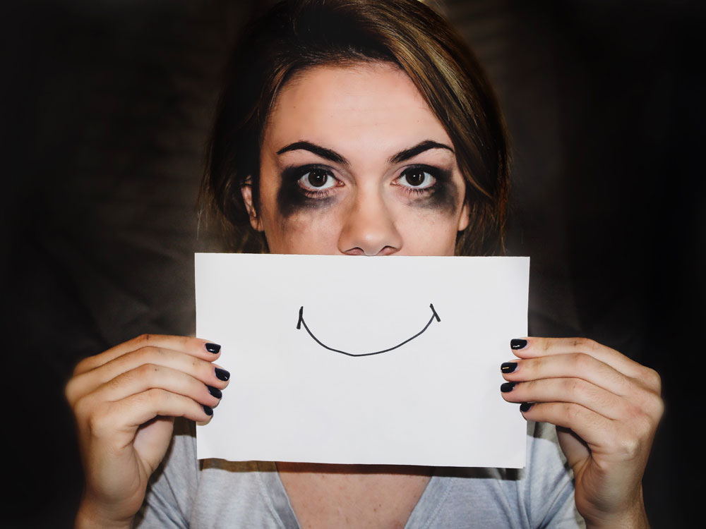 Une femme dont le maquillage a coulé cache le bas de son visage avec une feuille sur laquelle est dessiné un sourire